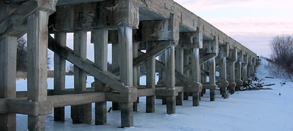 A crumbling railroad bridge over snow, the uprights are worn and look ready to give way.