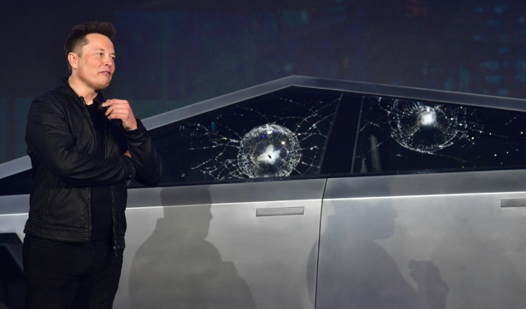 Elon musk next to two different "smashproof" windows on his cybertruck that he smashed.